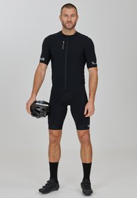 Schwarzes Radtrikot und Shorts mit kurzen Ärmeln, ausgestattet mit einem Reißverschluss vorn. Hält einen Fahrradhelm in einer Hand, trägt schwarze Radschuhe und Socken.