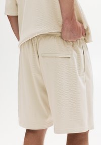 Un short en maille beige avec une taille élastique, des poches latérales et une poche arrière avec fermeture éclair. Le tissu a une texture respirante.