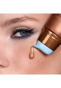 Bronse krem i en squeeze-tube med en lyseblå kork, som påfører en medium brun væske på huden. Nærbilde av hudtekstur og øyemakeup.