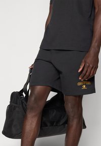 Μαύρα αθλητικά σορτς με κεντημένο κίτρινο λογότυπο "Champion", σε συνδυασμό με μονόχρωμο μαύρο t-shirt και μαύρη τσάντα duffel.