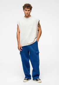 Grijze mouwloze top, ruime pasvorm; blauwe cargobroek met zijkanten zakken; zwart-witte sneakers; gestructureerde stof; effen achtergrond.
