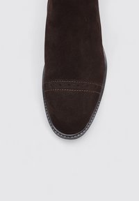 Chaussure en daim marron avec un bout arrondi, dotée de coutures décoratives et d'une semelle extérieure en caoutchouc noir texturé. Design minimaliste, sans ornementation.