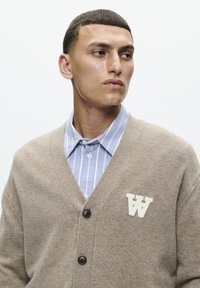 Beige strikket cardigan med V-udskæring, med mørke knapper og et hvidt broderet "AA" logo på venstre bryst, lagdelt over en blåstribet skjorte.