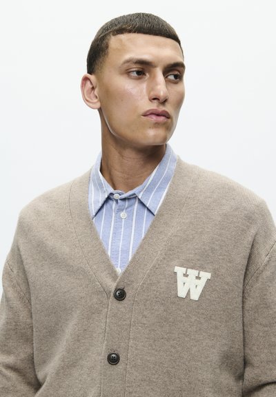Beige knitted cardigan with a V-neck, featuring dark buttons and a white embroidered "AA" logo on the left chest, layered over a blue striped shirt.
