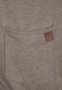 Lichtbruine gebreide stof met een voorzak met geribde randen en een klein bruin label met tekst. De textuur lijkt glad en consistent.