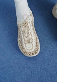Baskets à lacets avec des détails en tressage beige, une tige en maille blanche et une semelle en caoutchouc. Présente un logo vert sur la zone des orteils. Chaussettes blanches visibles.