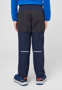 Pantaloni impermeabili blu navy con un pannello nero nella parte superiore, dettagli riflettenti e polsini elastici. Abbinati a scarpe da ginnastica blu e grigie.