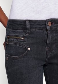 Vaqueros de mezclilla oscura con un ajuste slim, que presentan un bolsillo con cremallera, costuras en rojo contrastante y un acento metálico en la cinturilla.