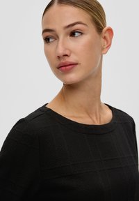 Czarna teksturowana bluzka z okrągłym dekoltem, z siatkowym wzorem. Tkanina wydaje się gruba i strukturalna, tworząc wizualną głębię.