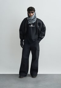 Zwarte gewatteerde bomberjack, zwarte t-shirt met logo, losse zwarte broek, grijze sjaal, zwarte handschoenen en zonnebril. Glad textuur, oversized pasvorm.