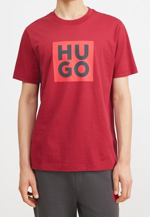 T-shirt en coton rouge avec un motif carré affichant le texte "HUGO" en lettres noires épaisses centré sur le devant. Manches courtes, coupe décontractée.