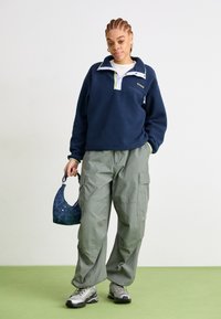 Pullover in pile blu navale con collare bianco e verde neon, abbinato a pantaloni cargo verde chiaro e sneakers argentate. Tiene in mano una borsa a motivo blu scuro.