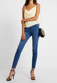 Top amarillo de satén con cuello drapeado y tirantes finos, combinado con jeans ajustados azules y tacones negros con tiras, complementado por un pequeño bolso adornado.