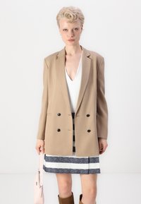 Beige kaksirivinen bleiseri sileässä kankaassa, yhdistettynä valkoiseen V-kaula-aukkopaitaan ja kuvioituun hameeseen. Pitelee pientä vaaleanpunaista käsilaukkua.