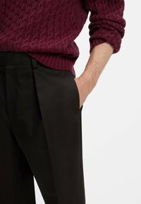 Il maglione a coste bordeaux presenta polsini a coste ed è indossato sopra pantaloni neri plissettati con tasche laterali, mostrando una texture liscia.