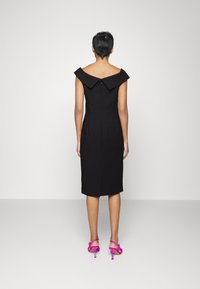 Schwarzes schulterfreies Kleid mit taillierter Silhouette, knielang, mit Reißverschluss auf der Rückseite und einem Schlitz. Kombiniert mit pinken, trippelnden High Heel Sandalen.