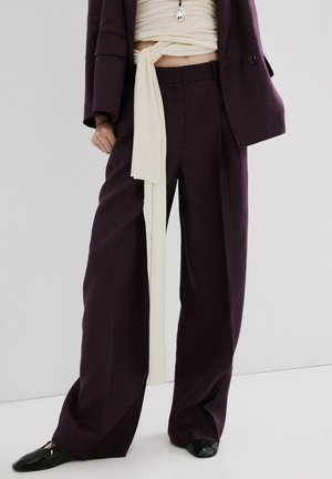 Personne portant un pantalon large violet foncé, un haut court croisé crème, une veste assortie et des ballerines noires sur un fond blanc.