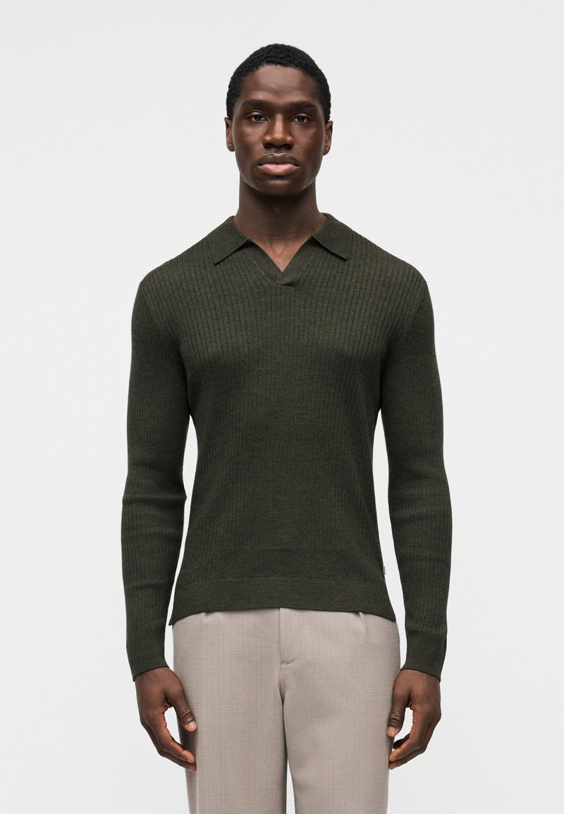 Maglione lavorato a maglia a coste di colore verde scuro con scollo a V e colletto corto, caratterizzato da maniche lunghe e design aderente. Indossato con pantaloni chiari.