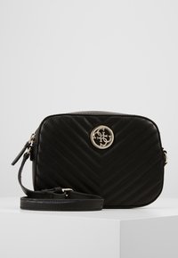 Sort quiltet crossover taske med justerbar rem og rundt guldlogo-emblem foran, præsenteret på hvidt piedestal.