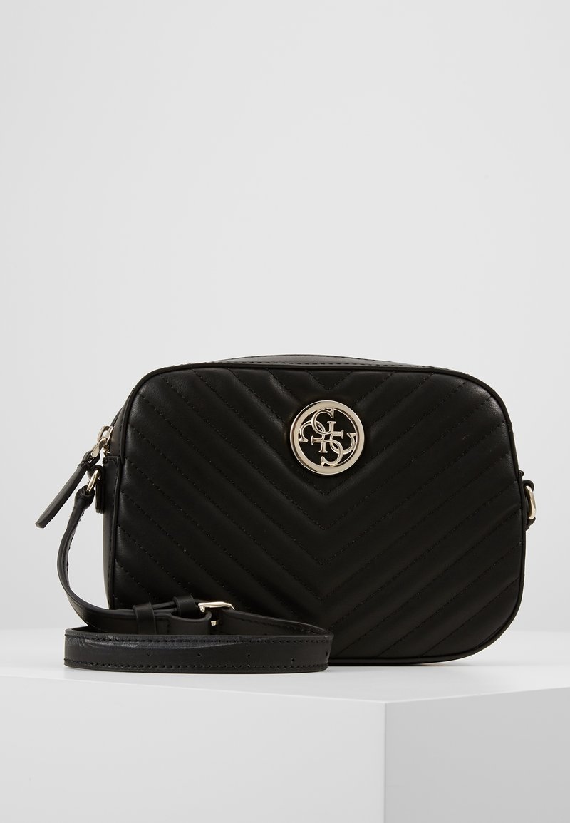 Sort quiltet crossover taske med justerbar rem og rundt guldlogo-emblem foran, præsenteret på hvidt piedestal.