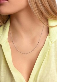 Sølv kædetsmykke med små runde accenter, hvilende på en halsudskæring med en synlig lysegul linnedbluse.
