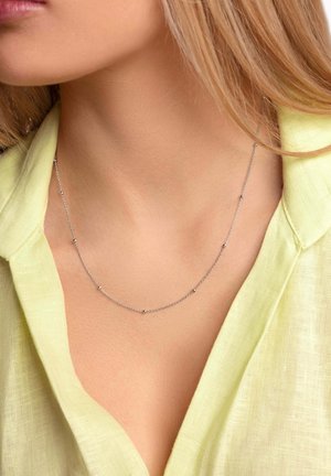 Collana in argento con piccoli accenti rotondi, poggiata su un décolleté con una canotta di lino giallo chiaro.