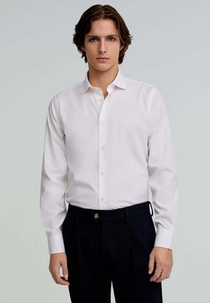 Camisa blanca de botones con mangas largas, que presenta un cuello clásico y una textura suave, combinada con pantalones oscuros.
