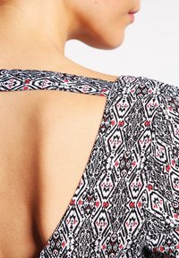 Black and white patterned fabric with red accents, featuring a crisscross back design and soft texture. Close-up view of the shoulder area.