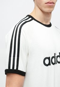 Biela tričko s čiernym lemom a tromi čiernymi pruhmi na rukáve; na prednej strane sa nachádza veľké čierne logo "adidas".