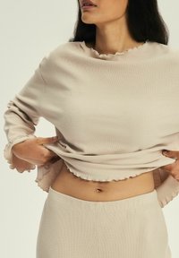 Beżowy top z prążkowanej tkaniny z falbaniastym dekoltem i mankietami, w połączeniu z pasującymi beżowymi spodniami, prezentujący prosty i teksturowany design.