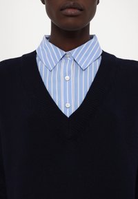 Gros plan d'une personne portant une chemise à rayures bleues et blanches sous un pull noir à col en V.