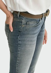 Tamsiai pilkos džinsai, turintys siaurą siluetą, priekinės kišenės ir rudos odinės diržą su auksinėmis detalėmis. Medžiaga yra šiek tiek tekstūruota.