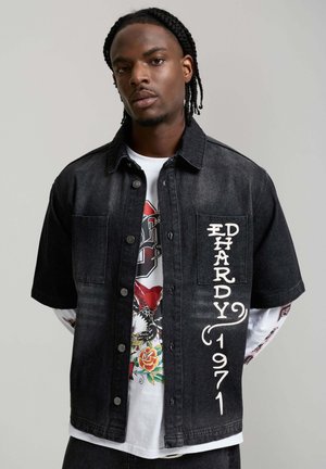 Uomo con capelli intrecciati che indossa una camicia di jeans nera a maniche corte con la scritta "ED HARDY 1971" sopra una maglia bianca a maniche lunghe con grafica.