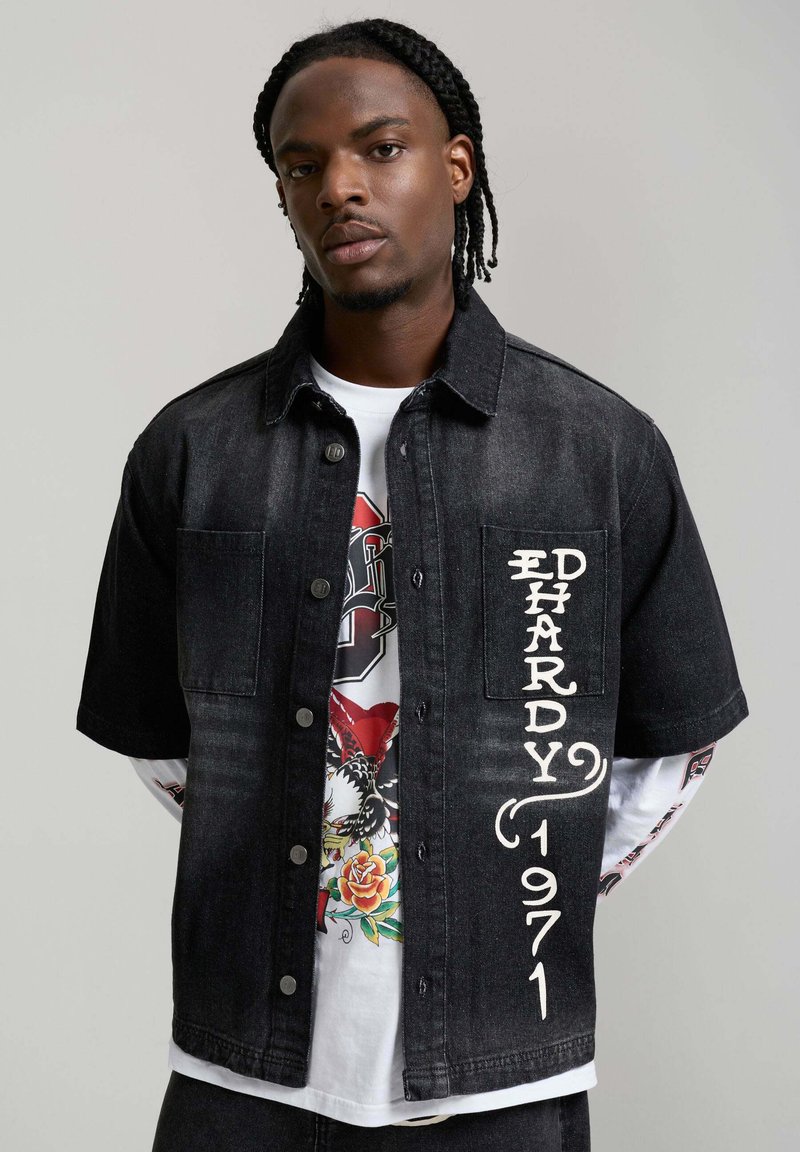 Mann mit geflochtenem Haar, trägt ein schwarzes kurzärmeliges Jeanshemd mit der Aufschrift "ED HARDY 1971" über einem weißen, langärmeligen T-Shirt mit Grafik.