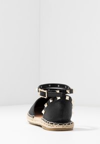 Espadrille plana de cuero negro con tachuelas doradas en forma de pirámide y hebilla en la correa del tobillo, mostrada desde la parte posterior.