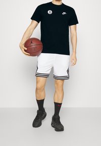 Camiseta negra de Nike con logo, pantalones cortos de baloncesto blancos con rayas negras, calcetines negros con detalles rojos y zapatillas deportivas negras, sosteniendo un balón de baloncesto.
