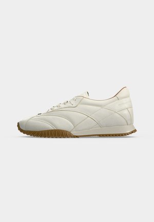 Sneaker bianca a taglio basso con dettagli di cuciture curve e suola in gomma naturale, mostrata di profilo su uno sfondo semplice.