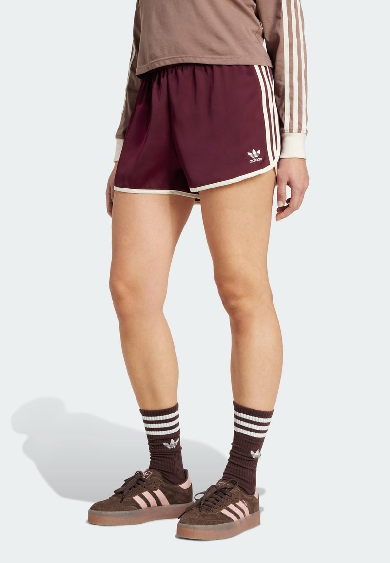 Pantalones cortos deportivos granate con ribetes blancos, con una cinturilla elástica y un logotipo bordado. Combinados con zapatillas marrones y calcetas a rayas.