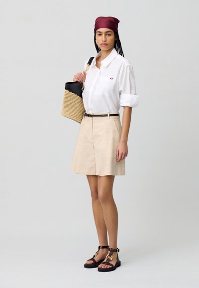 Camicia bianca con bottoni e un piccolo logo rosso, abbinata a pantaloni corti beige. Sandali marroni con dettagli dorati. Borsa intrecciata con fodera nera.