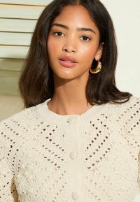 Beige pletený kardigan s texturovaným vzorem, žebrovaným výstřihem a předními knoflíky. Modelka má na sobě zlaté kruhové náušnice a jemný, přirozený makeup.