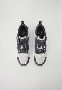 Sneakers with a gray and white mesh upper, black laces, and a patterned sole. Features a logo on the tongue and contrasting toe cap.