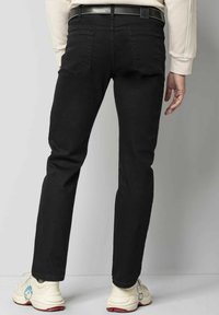 Jean en denim noir avec une coupe slim, disposant de deux poches arrière, de coutures décoratives et d'une étiquette à la taille. Porté avec des baskets blanches sur un fond gris.