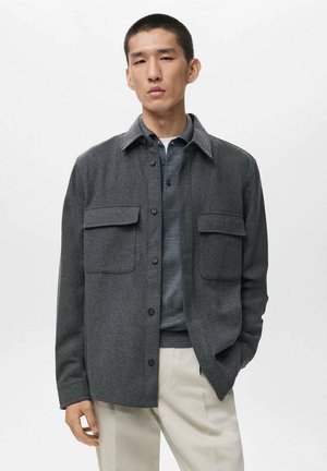 Dark gray wool-blend shirt jacket with a button front, two chest pockets, and a classic collar, paired with a navy sweater and light trousers.
