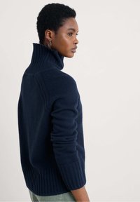 Temno modra turtleneck pulover iz mehkega materiala, z rebrastim teksturo na ovratniku in manšetah, ohlapnega kroja ter padlimi ramenskimi šivi.