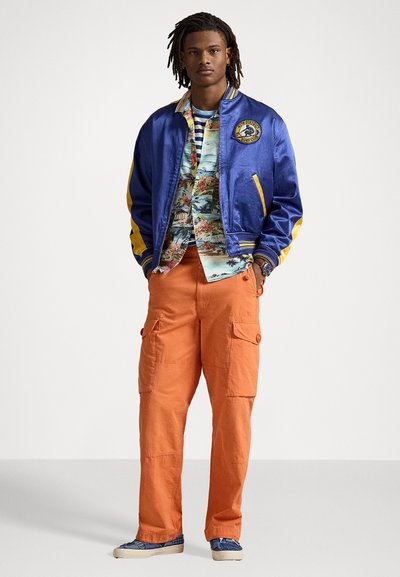 Blaues Satin-Bomberjackett mit gelben Akzenten und Logo, mehrfarbiges Blümchenhemd, orangefarbene Cargohose und blaue gemusterte Schuhe.