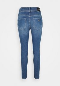 Skinny blue denim jeans with a mid-rise waist, featuring back pockets and a triangular logo patch on the waistband. Smooth texture.