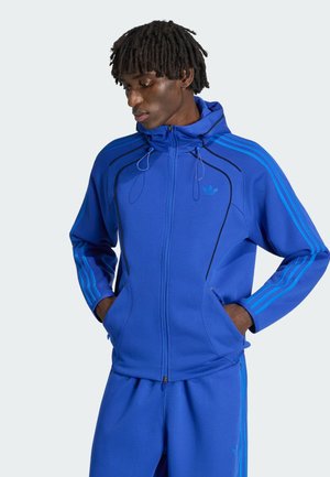 Młody mężczyzna z dredami, ubrany w niebieski dres Adidas z kurtką na zamek i rękami w kieszeniach, patrzący w dół na lewo.