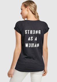 Sort T-shirt med kortærmer og afslappet pasform, der har fed hvid tekst på ryggen med teksten "STÆRKE SOM EN KVINDE." Glat stofstruktur.