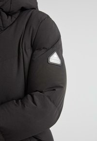 Sort quiltet vinterjakkeærme med et trekantet reflekterende "JACKIT" logo-patch på overarmen mod en grå baggrund.