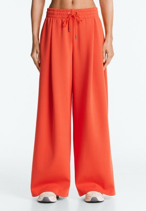 Wijde oranje broek met elastische tailleband en trekkoord, gedragen met beige sneakers door een staand persoon.